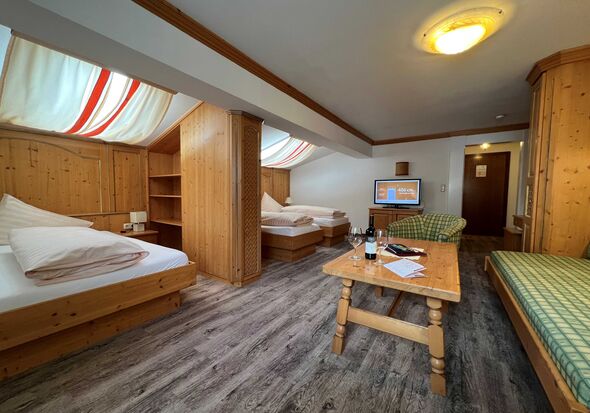 Zimmer mit Holzmöbeln, zwei Betten, Sofas, Tisch, Fernseher und sanfter Beleuchtung.